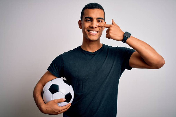 Knappe Afro-Amerikaanse man die voetballen houdt over een witte achtergrond Wijzend met de hand vinger naar gezicht en neus, vrolijk glimlachend. Schoonheidsconcept - Foto, afbeelding