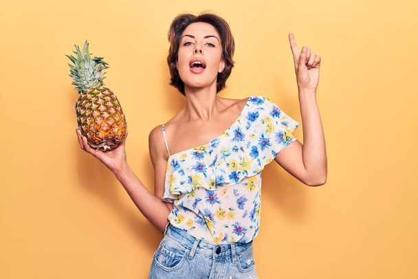 Young beautiful woman holding pineapple smiling with an idea or question pointing finger with happy face, number one  - Photo, Image