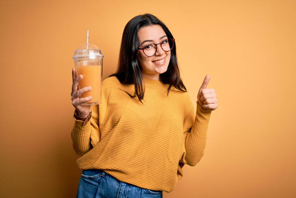 Young brunette woman drinking fresh orange juice from take away bottle over yellow background happy with big smile doing ok sign, thumb up with fingers, excellent sign - Photo, image