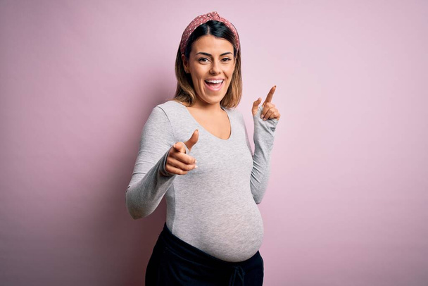 Fiatal gyönyörű barna nő terhes vár baba felett elszigetelt rózsaszín háttér mutató ujjak kamera boldog és vicces arcát. Jó energia és rezgések. - Fotó, kép