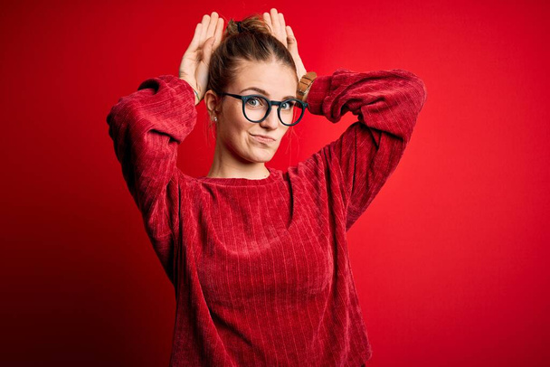 Giovane bella rossa donna indossa maglione casual su sfondo rosso isolato Fare coniglietto orecchie gesto con le mani palme cercando cinico e scettico. Concetto di coniglio pasquale. - Foto, immagini