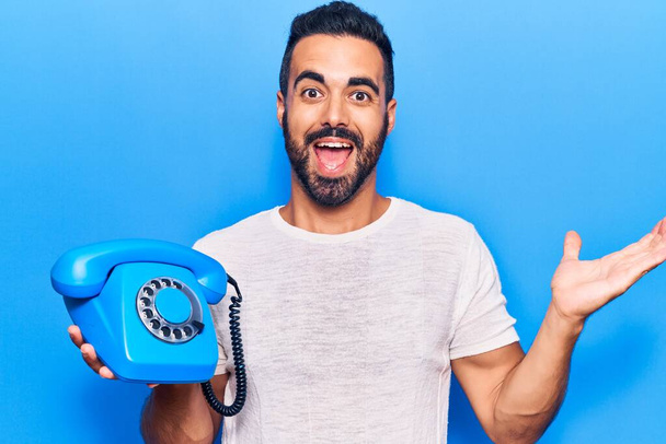 Jonge Spaanse man met vintage telefoon vieren prestatie met vrolijke glimlach en winnaar uitdrukking met opgeheven hand  - Foto, afbeelding