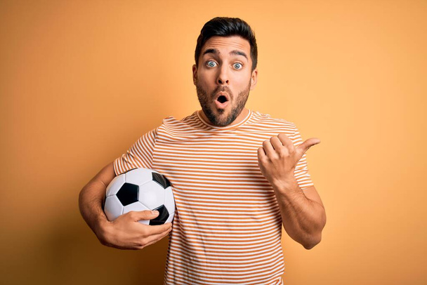 bell'uomo giocatore con la barba giocare a calcio tenendo pallone da calcio su sfondo giallo Sorpreso puntando con il dito della mano di lato, bocca aperta espressione stupita. - Foto, immagini