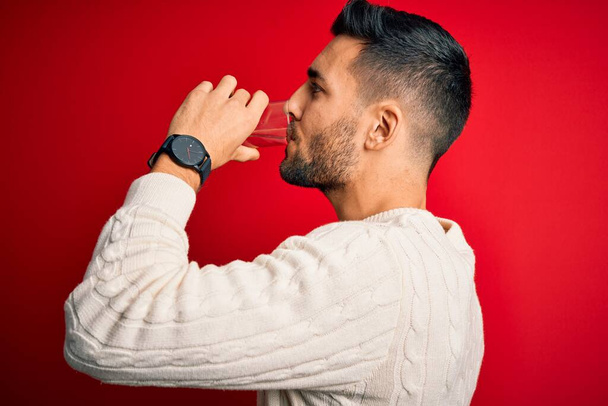 Young handsome man drinking glass of healthy water to refreshment standing over isolated red background - Foto, immagini