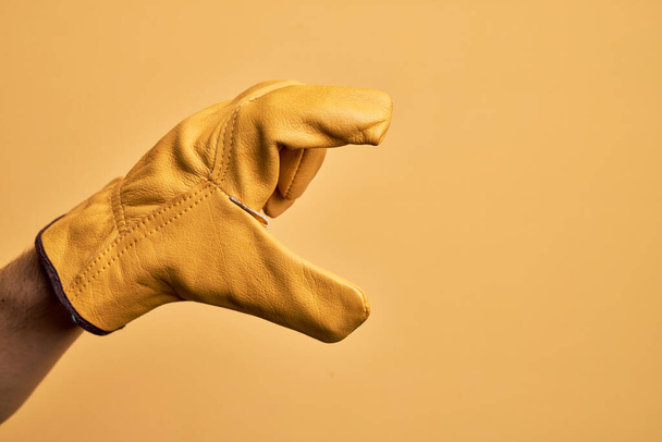 Hand of caucasian young man with gardener glove over isolated yellow background picking and taking invisible thing, holding object with fingers showing space - Photo, image