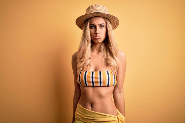 Jonge mooie blonde vrouw op vakantie dragen bikini en hoed over gele achtergrond depressief en zorgen te maken voor verdriet, huilen boos en bang. Droevige uitdrukking. - Foto, afbeelding