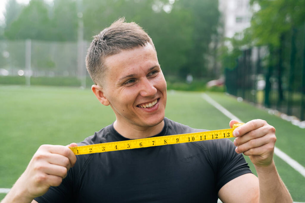 A close up of a cheerful male athlete is holding a centimeter tape in front of him. Muscle measurement concept - Photo, Image