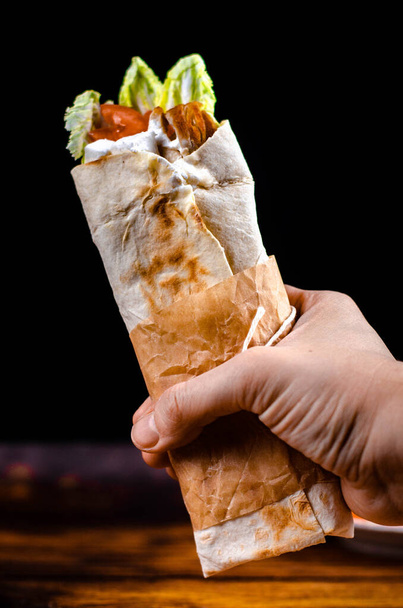 Shawarma turco o árabe con carne, verduras y salsa blanca en una mano sobre fondo negro
 - Foto, imagen