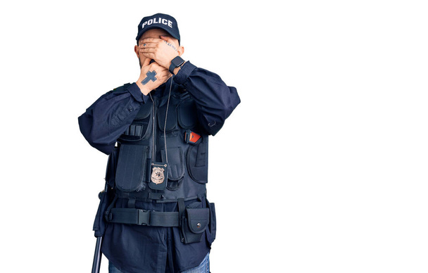 Young handsome man wearing police uniform covering eyes and mouth with hands, surprised and shocked. hiding emotion  - Foto, Imagen