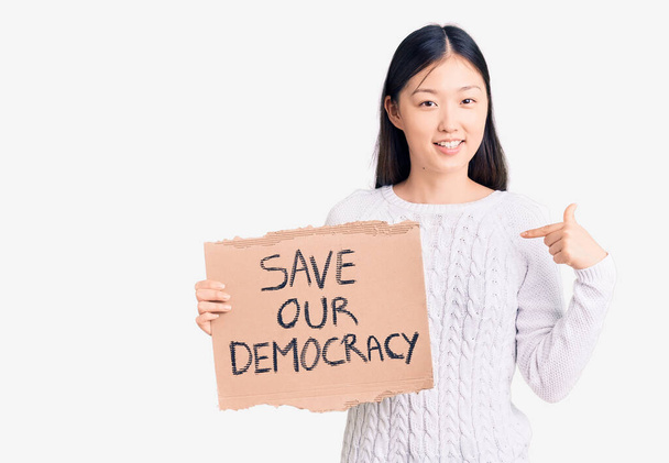 Молодая красивая китайская женщина держит сохранить нашу демократию картонный баннер указывая пальцем на один улыбается счастливый и гордый
  - Фото, изображение