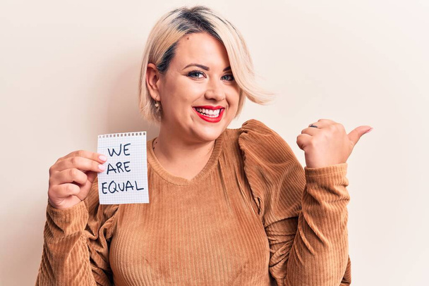 Beautiful blonde plus size woman asking for equality holding paper with we are equal message pointing thumb up to the side smiling happy with open mouth - Foto, immagini