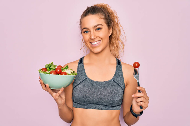 Молодая красивая блондинка спортивная женщина с голубыми глазами держит миску со здоровым салатом со счастливым лицом стоя и улыбаясь с уверенной улыбкой показывая зубы
 - Фото, изображение
