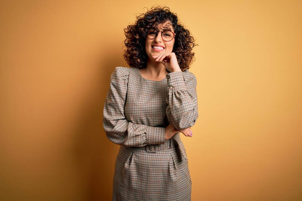 Beautiful arab business woman wearing dress and glasses standing over yellow background looking confident at the camera smiling with crossed arms and hand raised on chin. Thinking positive. - Foto, Imagem