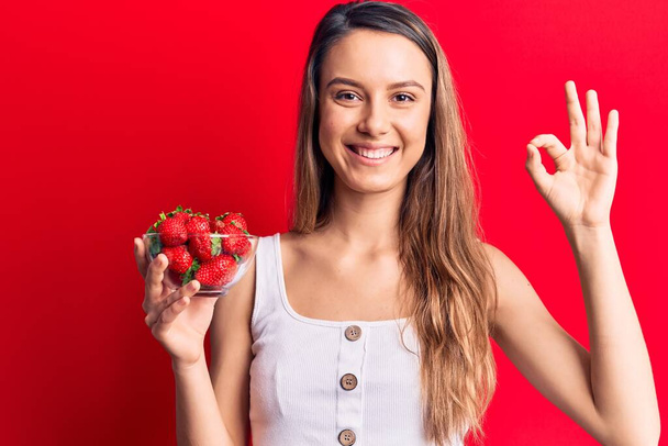 Junge schöne Mädchen hält Schale mit Erdbeeren tun ok Zeichen mit den Fingern, lächelt freundlich gestikulierend hervorragendes Symbol  - Foto, Bild