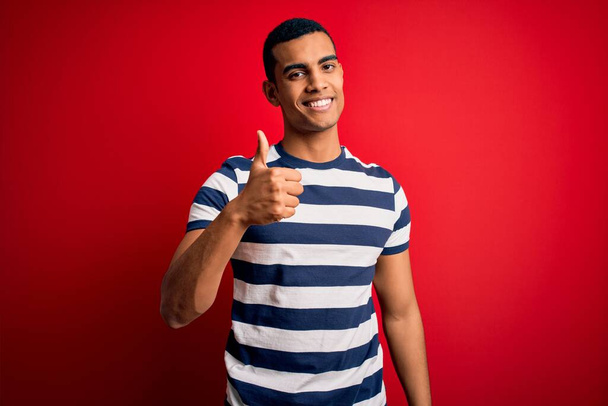 Красивый африканский американец в обычной полосатой футболке, стоящей на красном фоне и делающий счастливый жест вверх рукой. Утверждение выражения лица, смотрящего на камеру и показывающего успех
. - Фото, изображение
