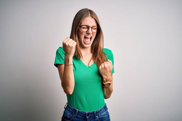 Young beautiful redhead woman wearing casual green t-shirt and glasses over white background celebrating surprised and amazed for success with arms raised and eyes closed. Winner concept. - Фото, зображення