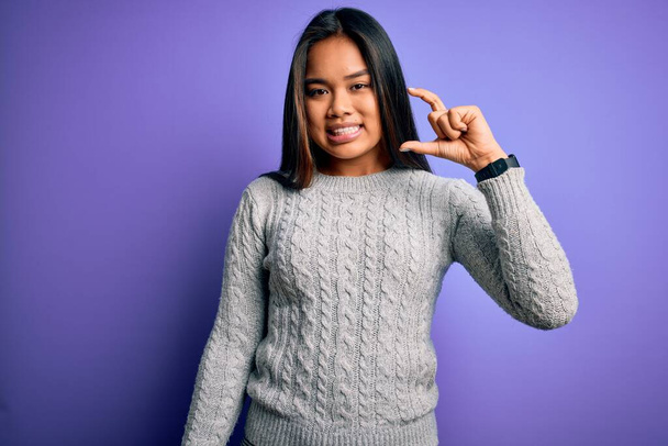 Young beautiful asian girl wearing casual sweater standing over isolated purple background smiling and confident gesturing with hand doing small size sign with fingers looking and the camera. Measure concept. - Photo, Image
