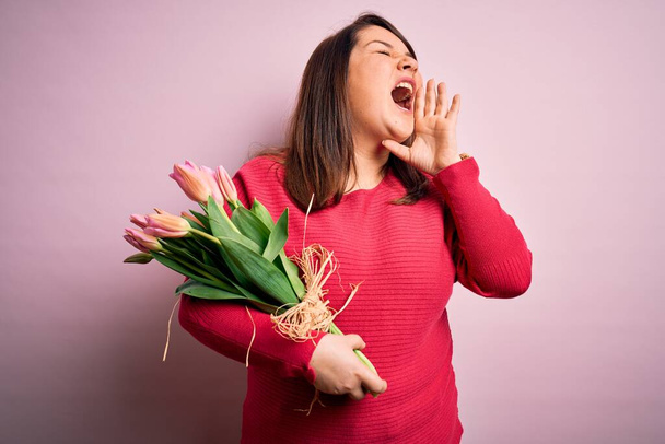 Belle femme taille plus tenant bouquet romantique de fleurs de tulipes naturelles sur fond rose criant et criant fort à côté avec la main sur la bouche. Concept de communication. - Photo, image