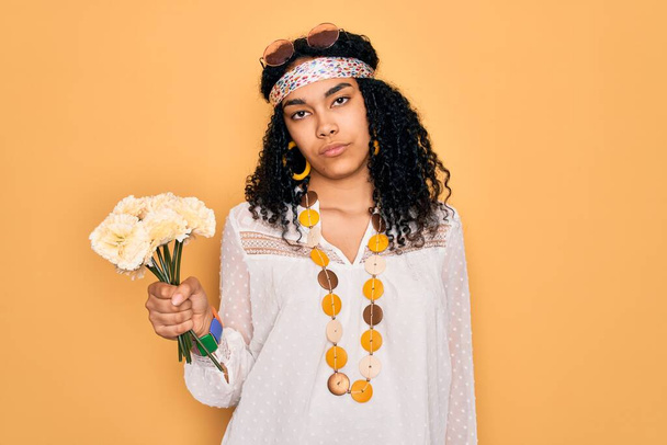 Jeune afro-américaine bouclée hippie femme portant des lunettes de soleil tenant bouquet de fleurs avec une expression confiante sur la pensée intelligente visage grave - Photo, image