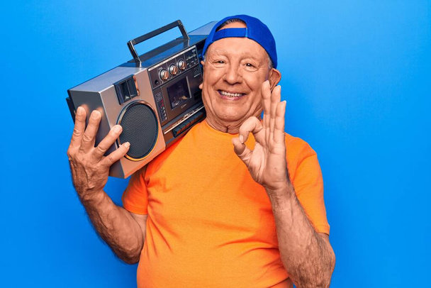 Senior moderní pohledný šedovlasý muž s čepicí poslouchající hudbu držící boombox dělá ok znamení s prsty, usmívající se přátelsky gestikulující vynikající symbol - Fotografie, Obrázek