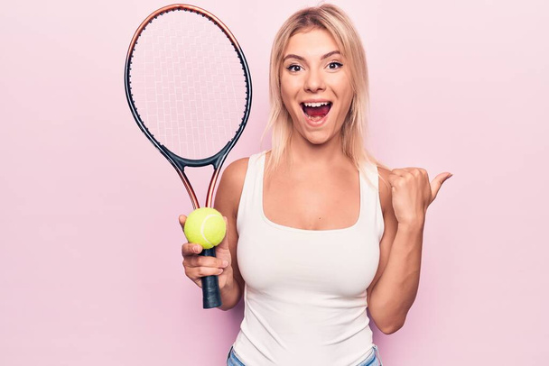 Młoda piękna blondynka sportowiec gra w tenisa za pomocą rakiety i piłkę na różowym tle wskazując kciuk do góry na bok uśmiechnięty szczęśliwy z otwartymi ustami - Zdjęcie, obraz