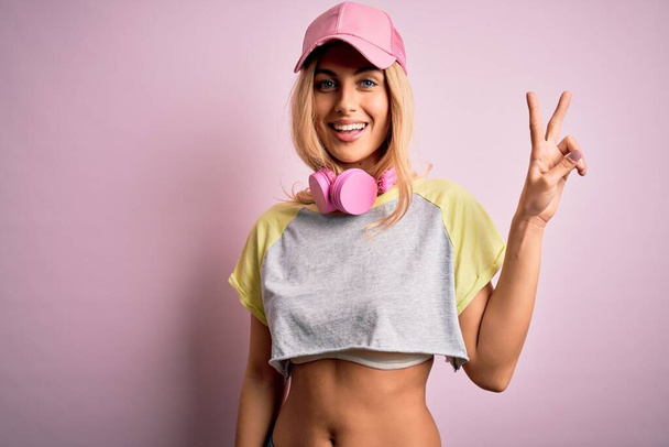 Young beautiful blonde sportswoman doing sport using headphones over pink background smiling with happy face winking at the camera doing victory sign. Number two. - 写真・画像
