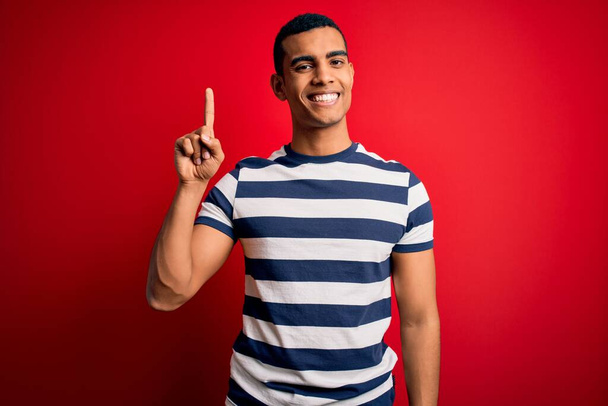 Schöner afrikanisch-amerikanischer Mann in lässigem gestreiftem T-Shirt, das über rotem Hintergrund steht und mit Finger Nummer eins zeigt, während er selbstbewusst und glücklich lächelt. - Foto, Bild