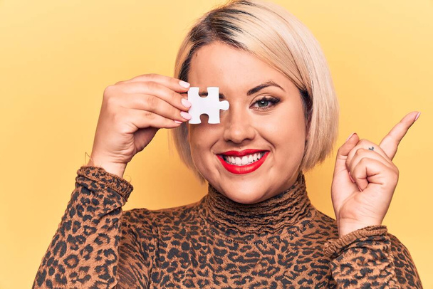 Young beautiful blonde plus size woman holding one piece of puzzle over eye smiling happy pointing with hand and finger to the side - 写真・画像