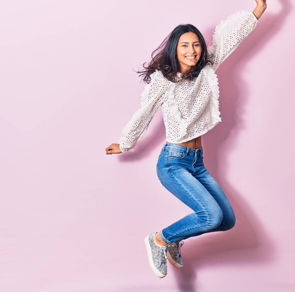 若い美しいラテン語の女性が幸せな笑顔カジュアルな服を着て。孤立したピンクの背景の上に顔に笑顔でジャンプ - 写真・画像