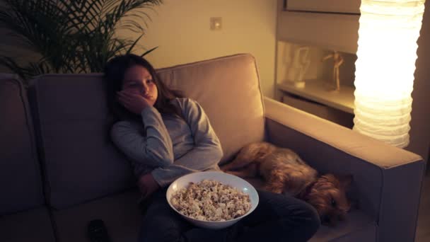 Tired girl sitting at home on the couch with her dog and watching a television. - Video, Çekim