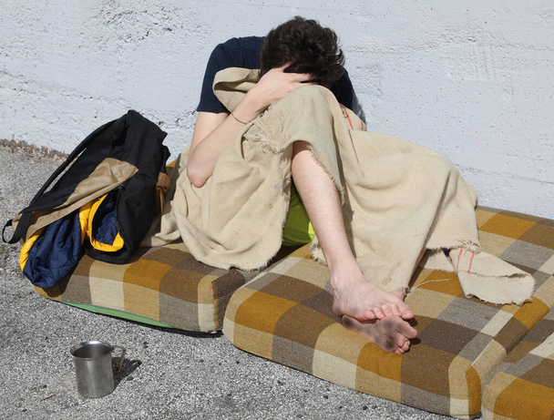 joven vagabundo se refugia bajo una manta sobre el colchón sucio en la acera de la ciudad
 - Foto, imagen