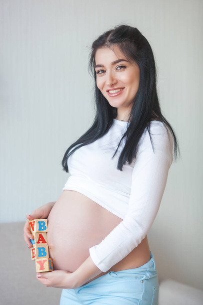 Attractive young woman expecting a child. Pregnant woman. Female indoors embracing her stomach. - Photo, Image