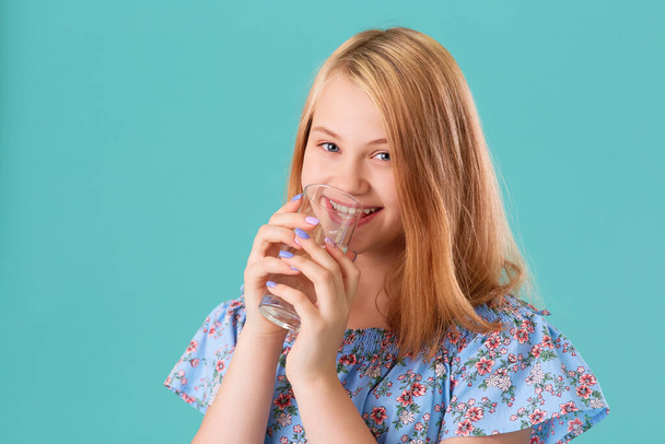 Portrait of a smiling girl drinking water from a glass glass. On a turquoise background - Foto, Bild