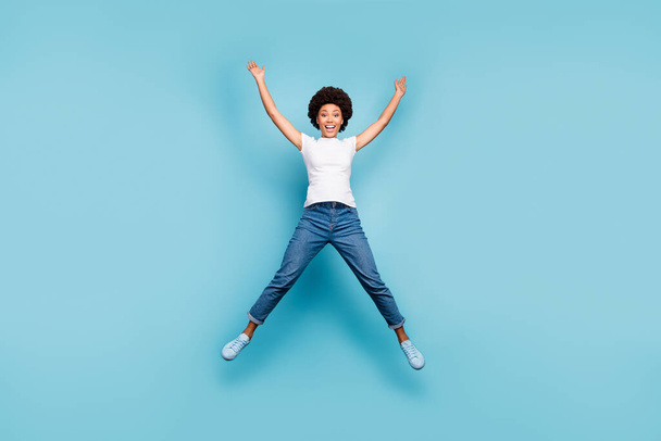 Full length photo of funny rejoicing dark skin wavy lady jumping high sunny summer day cheerful good mood wear casual white t-shirt jeans shoes isolated blue color background - Foto, Bild