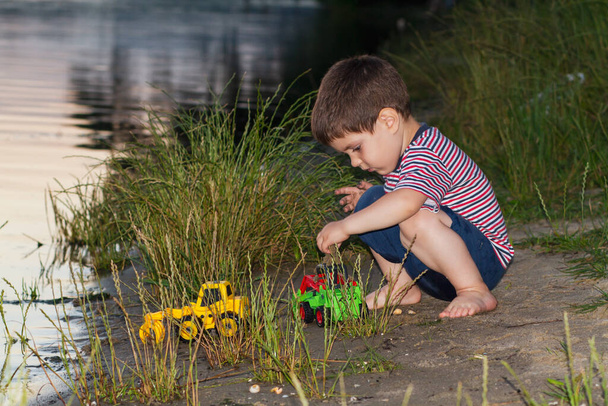 Ένα αγοράκι τριών ετών παίζει στην όχθη του ποταμού με φωτεινά παιχνίδια, σκάβει άμμο. Παιδικά παιχνίδια κοντά στη λίμνη στον καθαρό αέρα το καλοκαίρι - Φωτογραφία, εικόνα