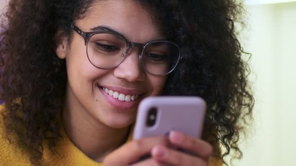 Close up view of Cheerful african attractive woman in eyeglasses using smartphone in living room - Záběry, video