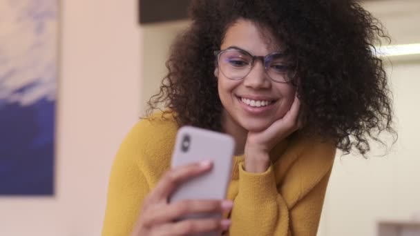 Jovem mulher africana atraente usando smartphone, em seguida, tornando-se surpresa e adiando seus óculos enquanto se senta na cadeira dentro de casa
 - Filmagem, Vídeo