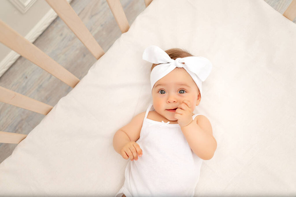 πορτρέτο ενός μικρού κοριτσιού 6 μηνών με φιόγκο στην κούνια στο παιδικό δωμάτιο με λευκά ρούχα και κοιτάζοντας την κάμερα, το πρωινό του μωρού, την έννοια των βρεφικών προϊόντων - Φωτογραφία, εικόνα
