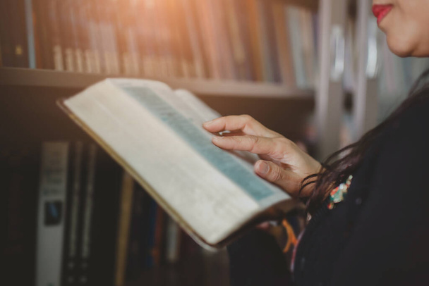 κλείσιμο των χεριών μιας γυναίκας κρατώντας και διαβάζοντας την Αγία Γραφή πάνω θολά βιβλία στη βιβλιοθήκη, επιλεκτική εστίαση στο χέρι της γυναίκας  - Φωτογραφία, εικόνα