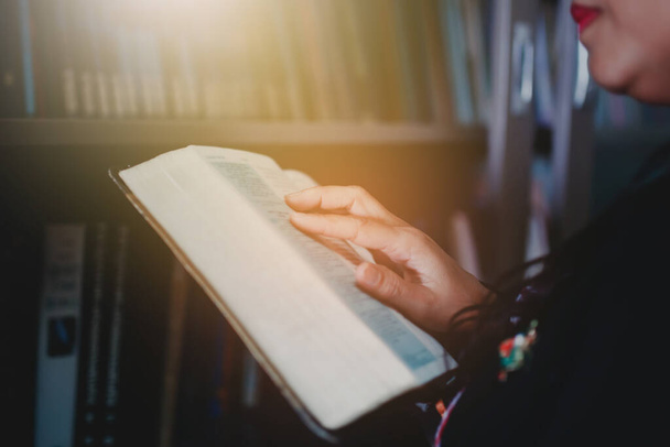Κλείστε τα χέρια μιας γυναίκας κρατώντας και διαβάζοντας την Αγία Γραφή πάνω από θολά βιβλία στο ράφι, επιλεκτική εστίαση στο χέρι της γυναίκας  - Φωτογραφία, εικόνα