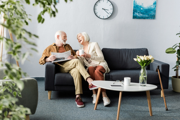 Selektiver Fokus glücklicher Senioren beim Lachen auf Sofa neben Couchtisch mit Tulpen - Foto, Bild