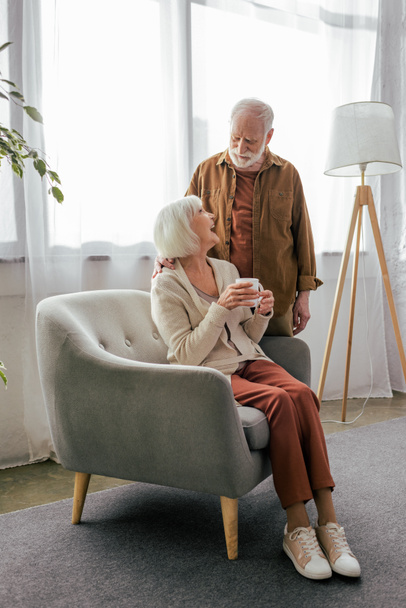 heureuse femme âgée assise dans un fauteuil avec une tasse de thé et parlant au mari - Photo, image