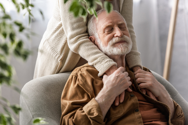 Ausgeschnittene Ansicht einer Frau, die Hände eines glücklichen älteren Mannes hält, der mit geschlossenen Augen sitzt - Foto, Bild