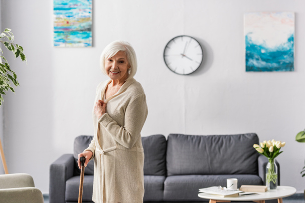 happy senior woman looking at camera while standing with walking stick at home - Foto, Bild