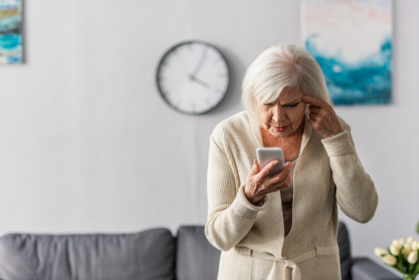 reflexiva mujer mayor tocando la cabeza mientras se utiliza el teléfono inteligente
 - Foto, imagen
