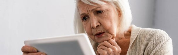horizontales Bild einer nachdenklichen Seniorin, die die Hand am Kinn hält, während sie ein digitales Tablet benutzt - Foto, Bild