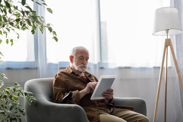 концентрированный пожилой человек с помощью цифрового планшета, сидя в кресле
 - Фото, изображение