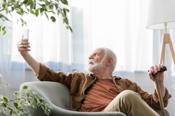 selective focus of senior man sitting in armchair and taking selfie on smartphone - Photo, Image