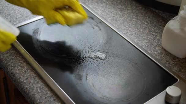 La mano de mujer en guantes de goma amarilla lava la estufa de inducción con detergente en aerosol
 - Imágenes, Vídeo