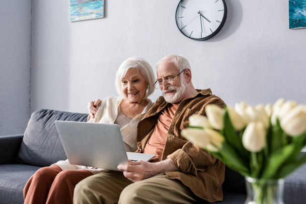 szelektív fókusz boldog idős pár ül a kanapén, és használja laptop közelében csokor tulipán - Fotó, kép
